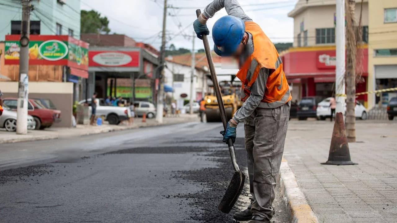 A road construction worker in a hard hat and orange vest applying fresh asphalt on a city street; the worker's face is blurred for privacy.