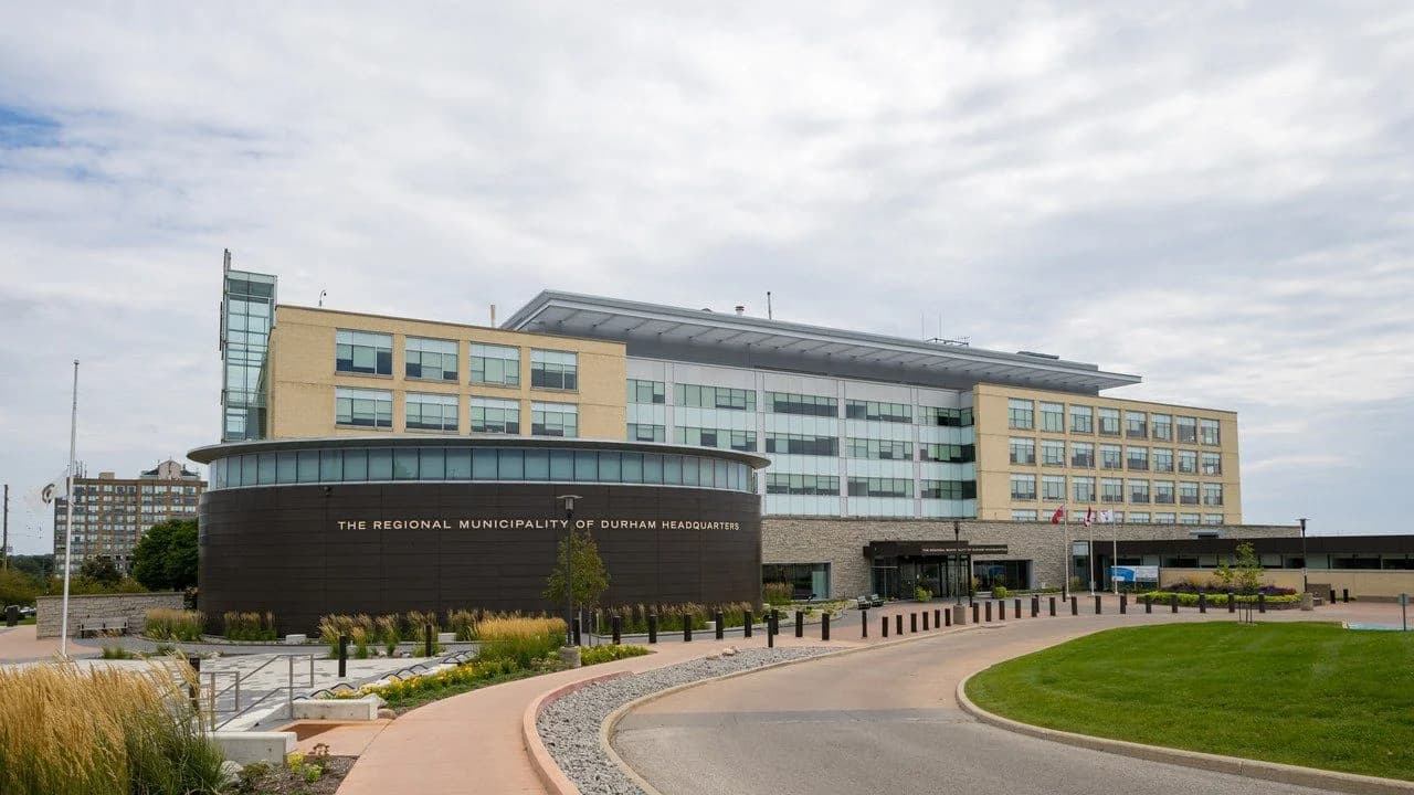 The Regional Municipality of Durham headquarters building in Whitby, Ontario — a modern tan-brick and glass office building with a curved dark entry rotunda, landscaped driveway and flags under a cloudy sky.
