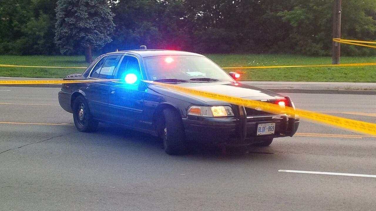 An unmarked police cruiser with emergency lights on, parked beside police tape on a road